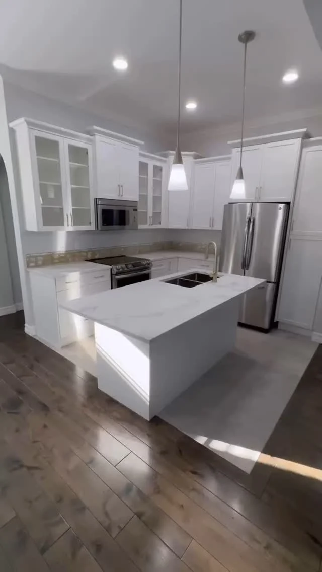 Bright white kitchen with island sink, gold hardware, glass-front upper cabinets, pendant lights and stainless appliances.