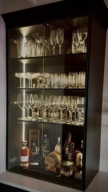 Dark glass-front bar cabinet displaying stemware, bottles and warm interior lighting.