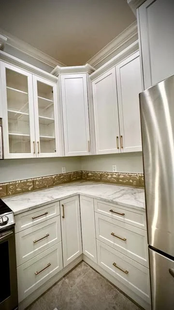 White corner kitchen cabinetry, glass-front upper cabinets, gold pulls and stainless refrigerator.