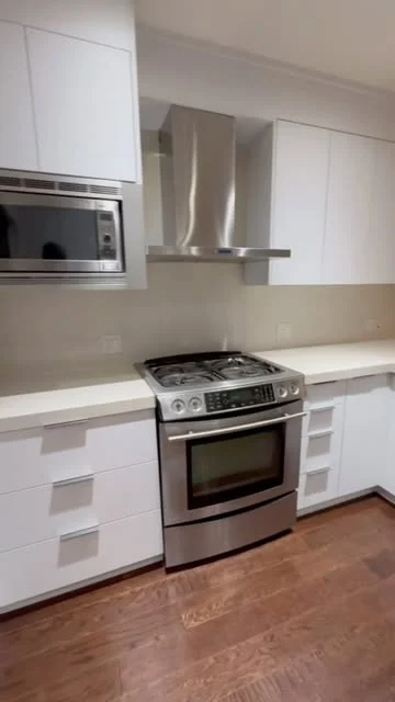 Modern white kitchen with flat cabinet fronts, stainless range and hood, pale backsplash and wood floor.