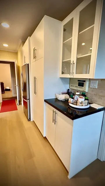 White kitchen pantry and glass-front wall cabinets above a black countertop, with light wood floor.